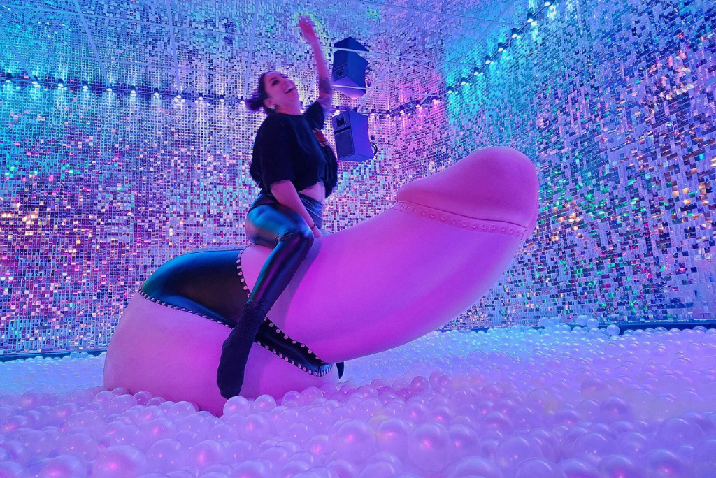 Person riding a vibrant mechanical penis in a colorful, illuminated setting with reflective walls and soft bubble flooring, embodying lively entertainment for parties and events.