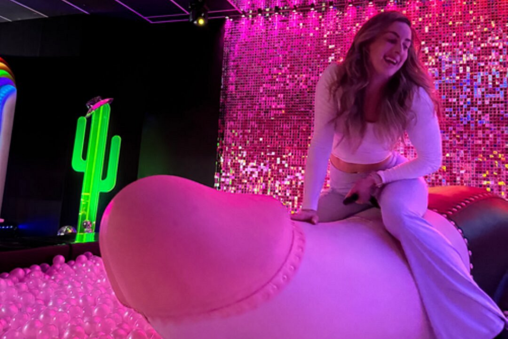Woman joyfully riding a mechanical penis attraction, surrounded by vibrant pink lights, a green cactus decoration, and colorful party elements, embodying the fun and energetic atmosphere of events like drag shows and bachelorette parties.
