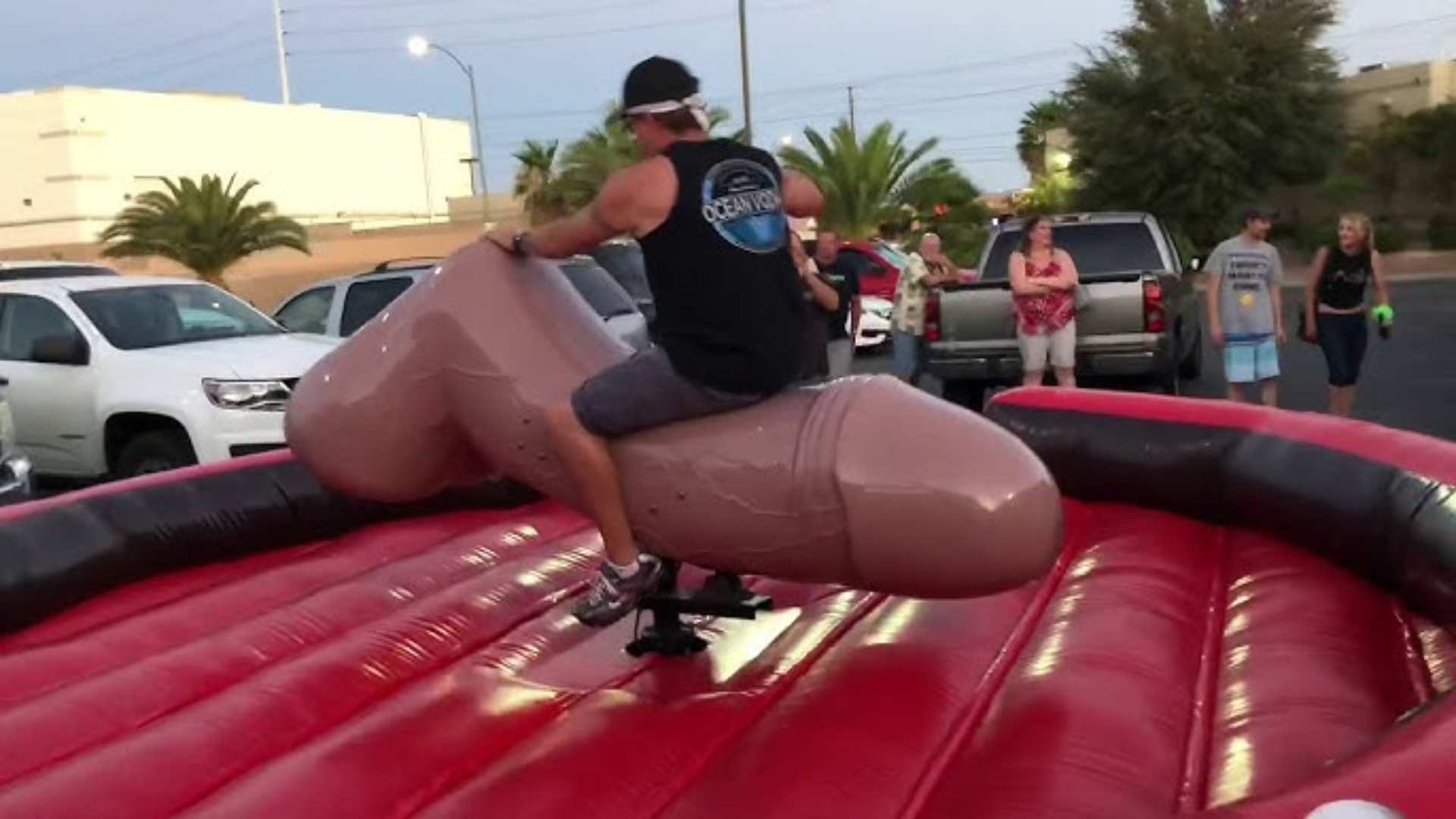 Man riding a mechanical penis on a red inflatable surface at a lively outdoor party, surrounded by amused onlookers, emphasizing bold celebration and outrageous fun.