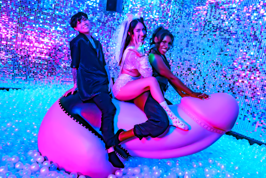 Three individuals enjoying a ride on a colorful mechanical penis attraction, surrounded by shimmering disco-style decorations and pearl-like balls, embodying fun and vibrant party energy.