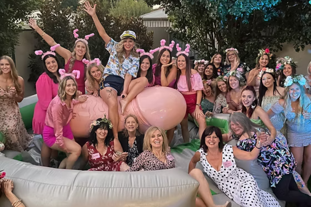 Group of women celebrating on a mechanical penis ride at a lively party, wearing fun outfits and accessories, surrounded by vibrant decorations and an energetic atmosphere.