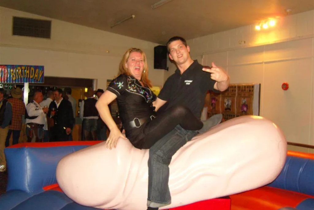 Woman riding a mechanical penis ride, smiling and posing playfully, with a man giving a peace sign beside her, in a festive party setting with a "Birthday" banner in the background.